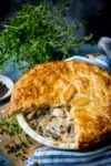 Chicken and mushroom pie with a slice removed on a wooden table with fresh herbs in the background