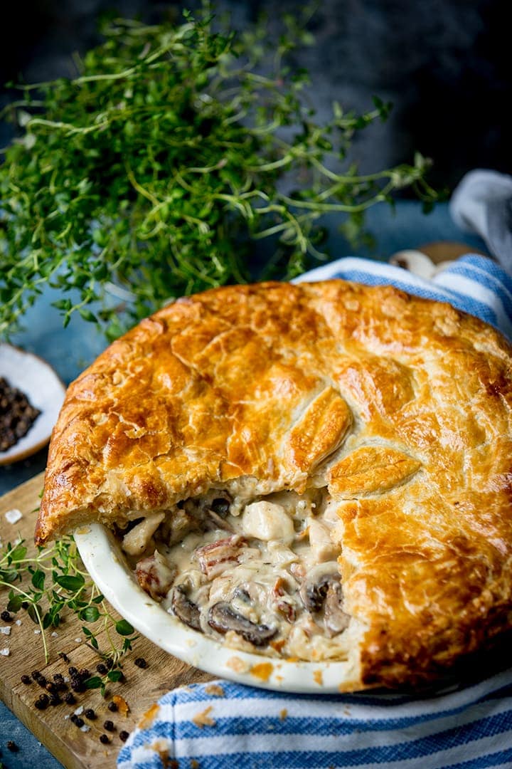Chicken and mushroom pie with a slice removed on a wooden table with fresh herbs in the background