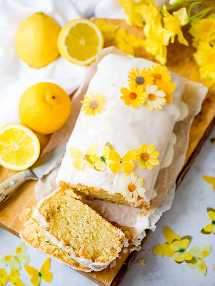 Overhead photo of vegan lemon drizzle cake with a slice taken out. Sugar flowers on top of the cake.