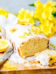 Sliced vegan lemon drizzle cake with white icing on yellow sugar flowers on a wooden board