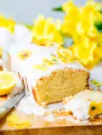 Sliced vegan lemon drizzle cake with white icing on yellow sugar flowers on a wooden board