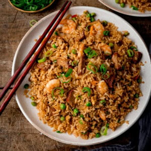 Top down square image of a white plate of Yeung Chow Fried Rice with prawns, pork, peas, and chopped spring onions, served with chopsticks. The plate is on a wooden table with a further plate of rice and a dish of chopped spring onions just slightly in shot.