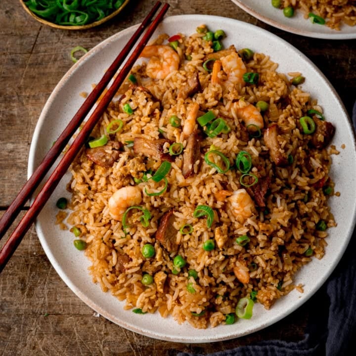 Top down square image of a white plate of Yeung Chow Fried Rice with prawns, pork, peas, and chopped spring onions, served with chopsticks. The plate is on a wooden table with a further plate of rice and a dish of chopped spring onions just slightly in shot.