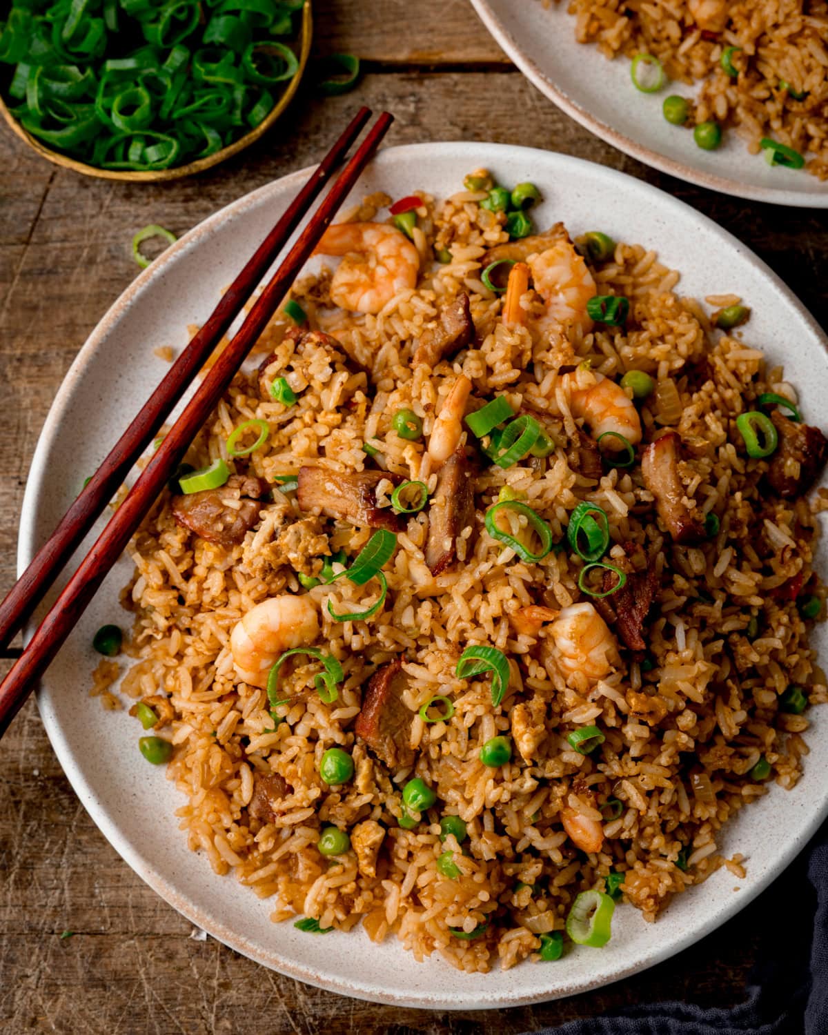 Top down image of a white plate of Yeung Chow Fried Rice with prawns, pork, peas, and chopped spring onions, served with chopsticks. The plate is on a wooden table next to a further plate of rice and a dish of chopped spring onions.