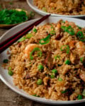 Side on image of a white plate of Yeung Chow Fried Rice with prawns, pork, peas, and chopped spring onions, served with chopsticks. The plate is on a wooden table next to a further plate of rice and a dish of chopped spring onions.