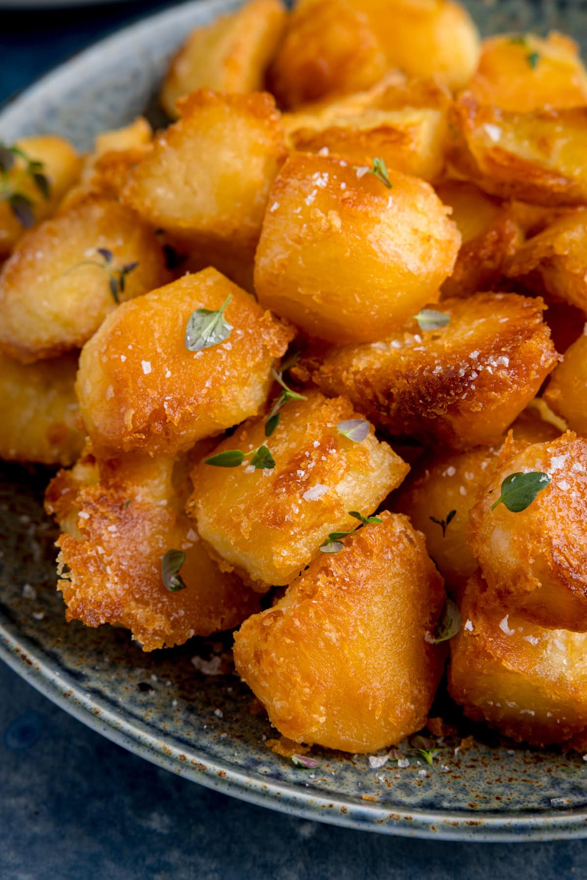 A close-up of golden, crispy perfect roast potatoes garnished with sea salt and fresh thyme on a speckled ceramic plate.