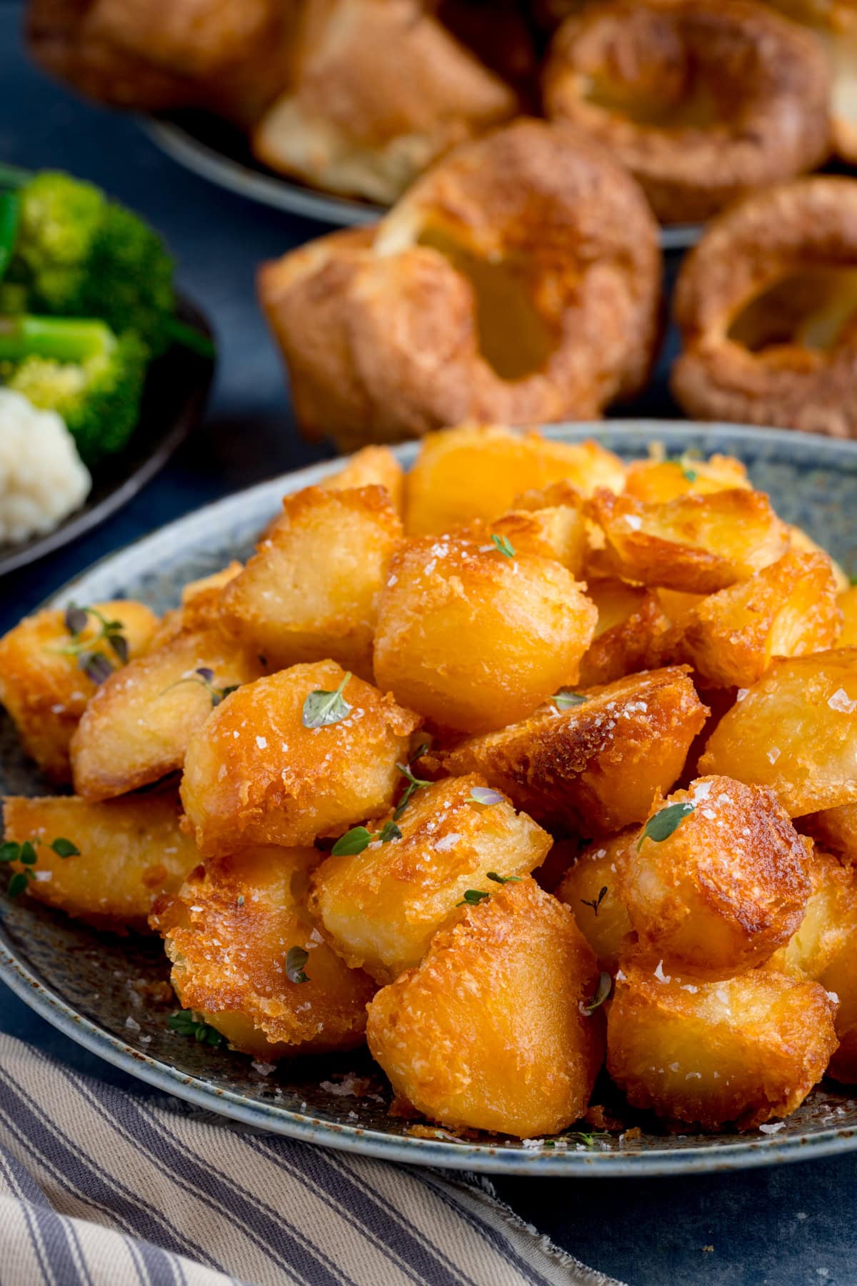 A plate of golden, crispy roast potatoes garnished with herbs, with Yorkshire puddings and vegetables visible in the background.