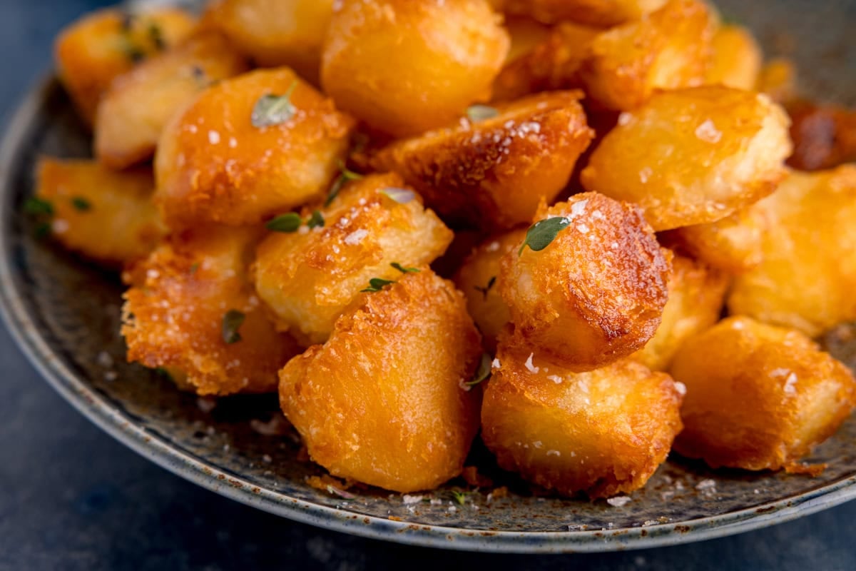 A wide image of a blue plate with golden, crispy roast potatoes garnished with sea salt flakes and fresh thyme.