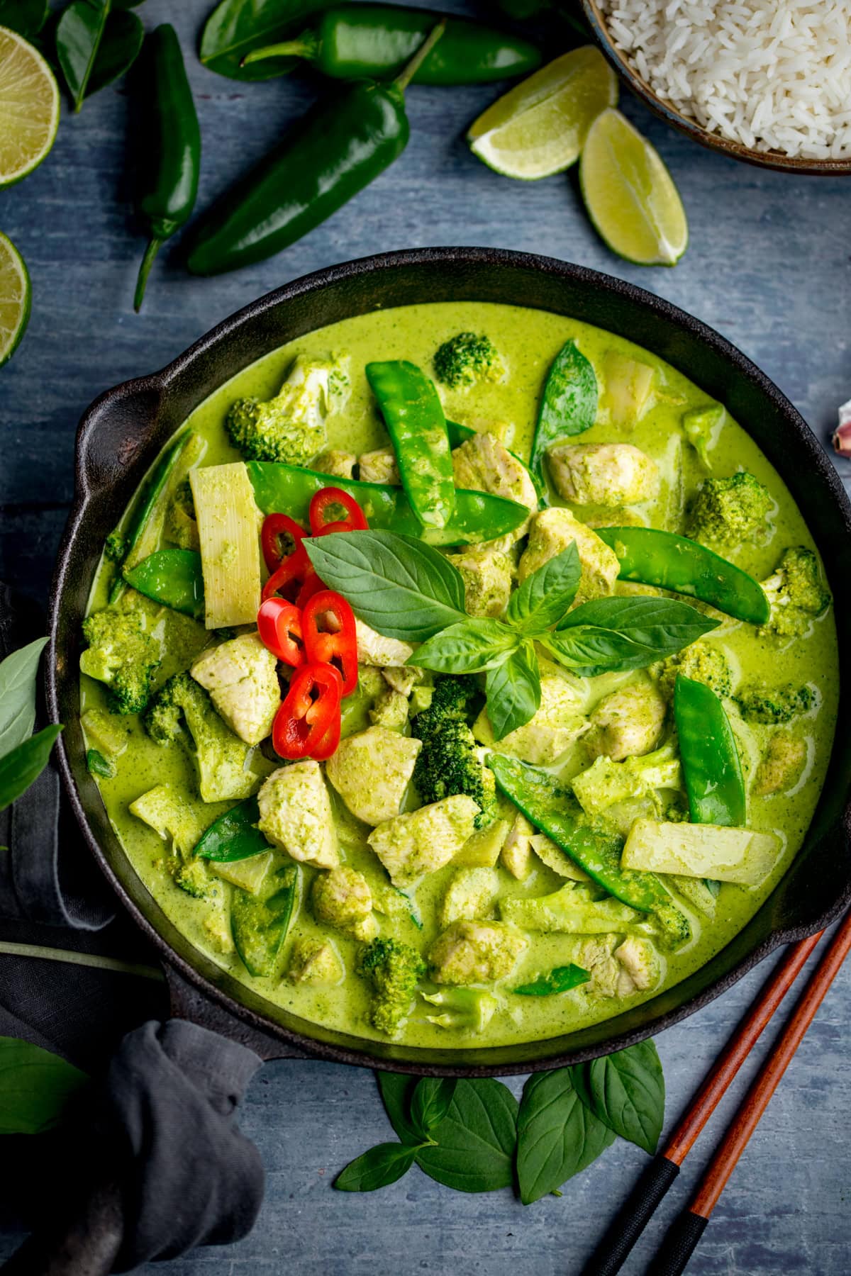 A bowl of Thai Green Curry made with authentic Thai green curry paste, featuring chicken, broccoli, bamboo shoots, mangetout, red chilli slices, and fresh basil, served with white rice and lime wedges.