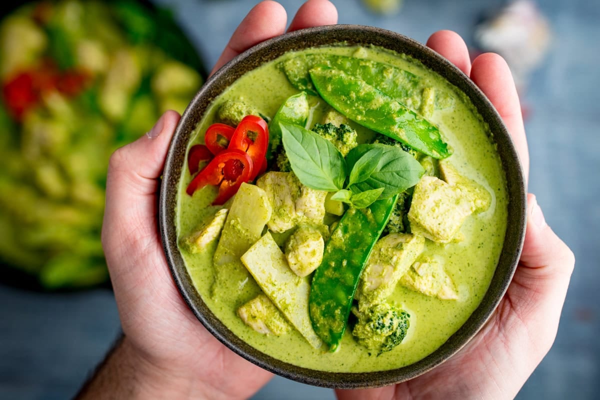 A bowl of Thai Green Curry with chicken, mangetout, broccoli, red chilli slices, and fresh basil leaves, held in two hands.