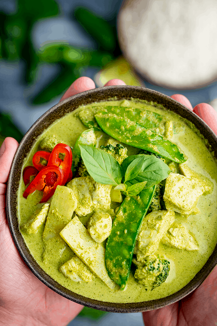 A bowl of Thai green chicken curry with vegetables being held in two hands