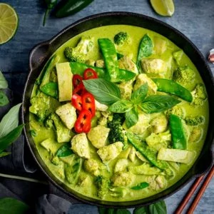 Square image of Pan of Thai green chicken curry with vegetables on blue background