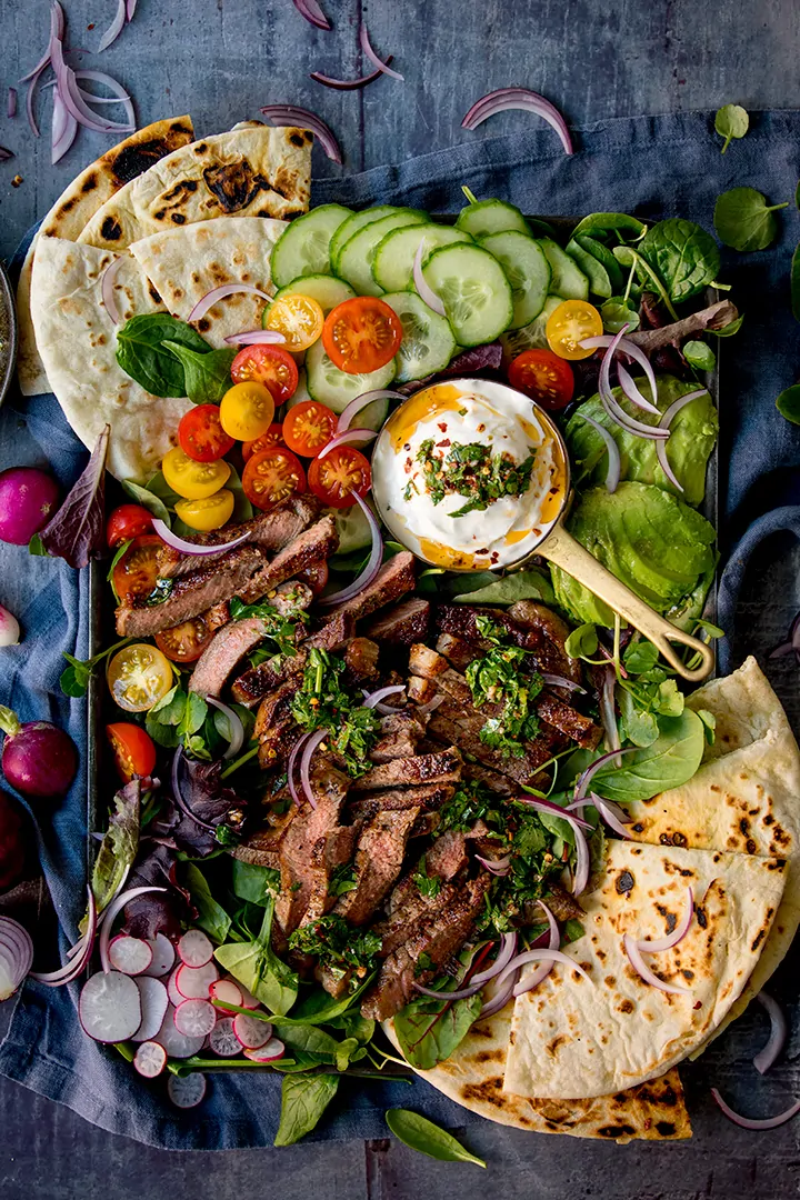 Overhead image of a large salad platter with strips of steak, chimichurri sauce and flatbreads on a blue background