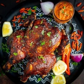 Roasted peri peri chicken on a tray next to a jar of homemade peri peri sauce. There are garnishes surrounding the chicken.