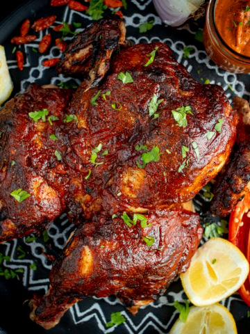 Roasted peri peri chicken on a tray next to a jar of homemade peri peri sauce. There are garnishes surrounding the chicken.