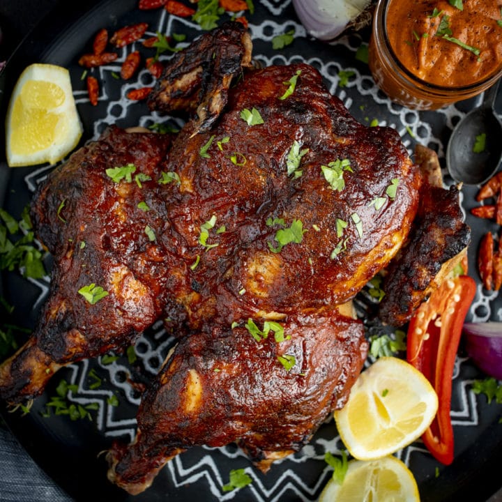 Roasted peri peri chicken on a tray next to a jar of homemade peri peri sauce. There are garnishes surrounding the chicken.