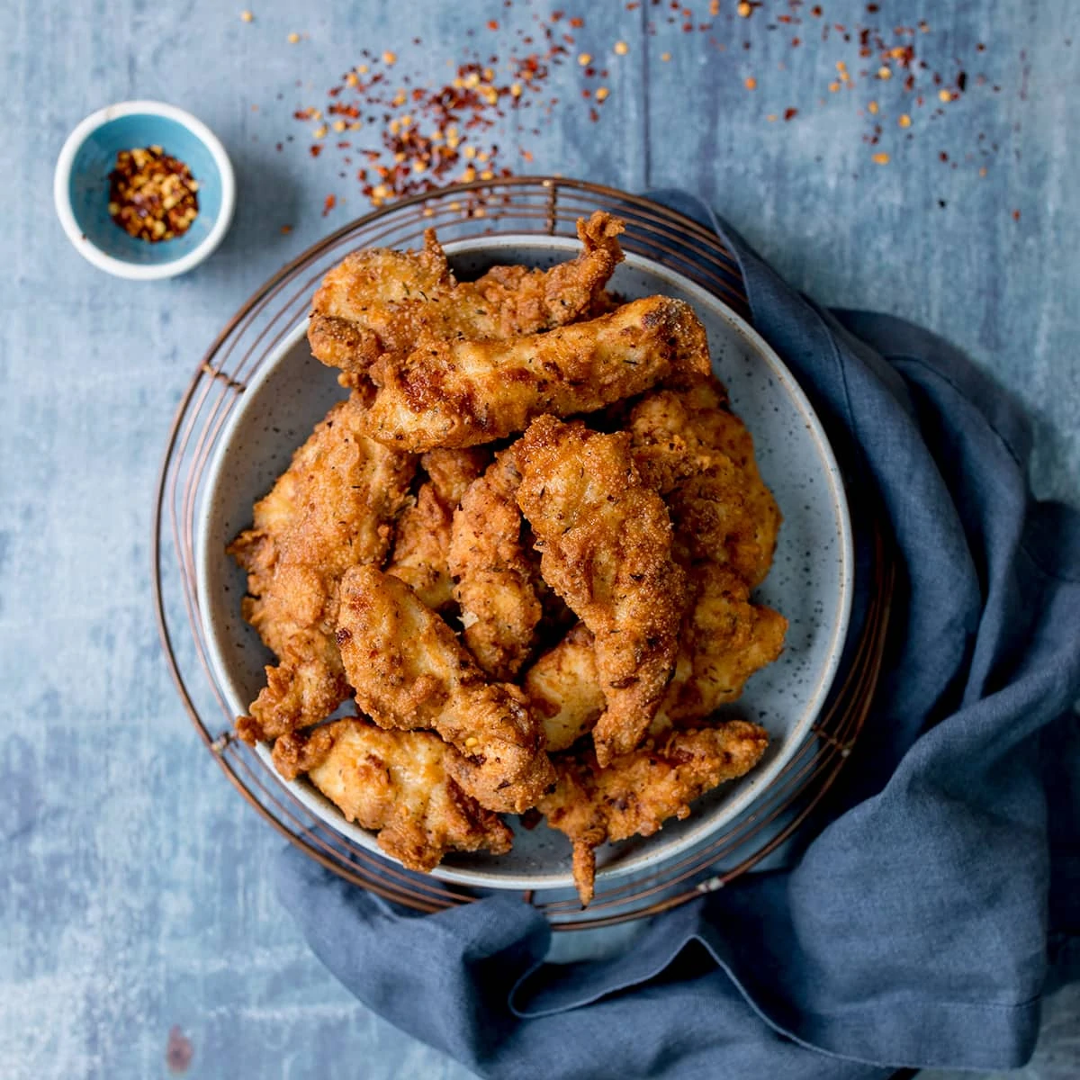 Overhead picture of Crispy Fried Buttermilk Chicken in a blue bowl with a small bowl of red pepper chilli flakes on a blue background