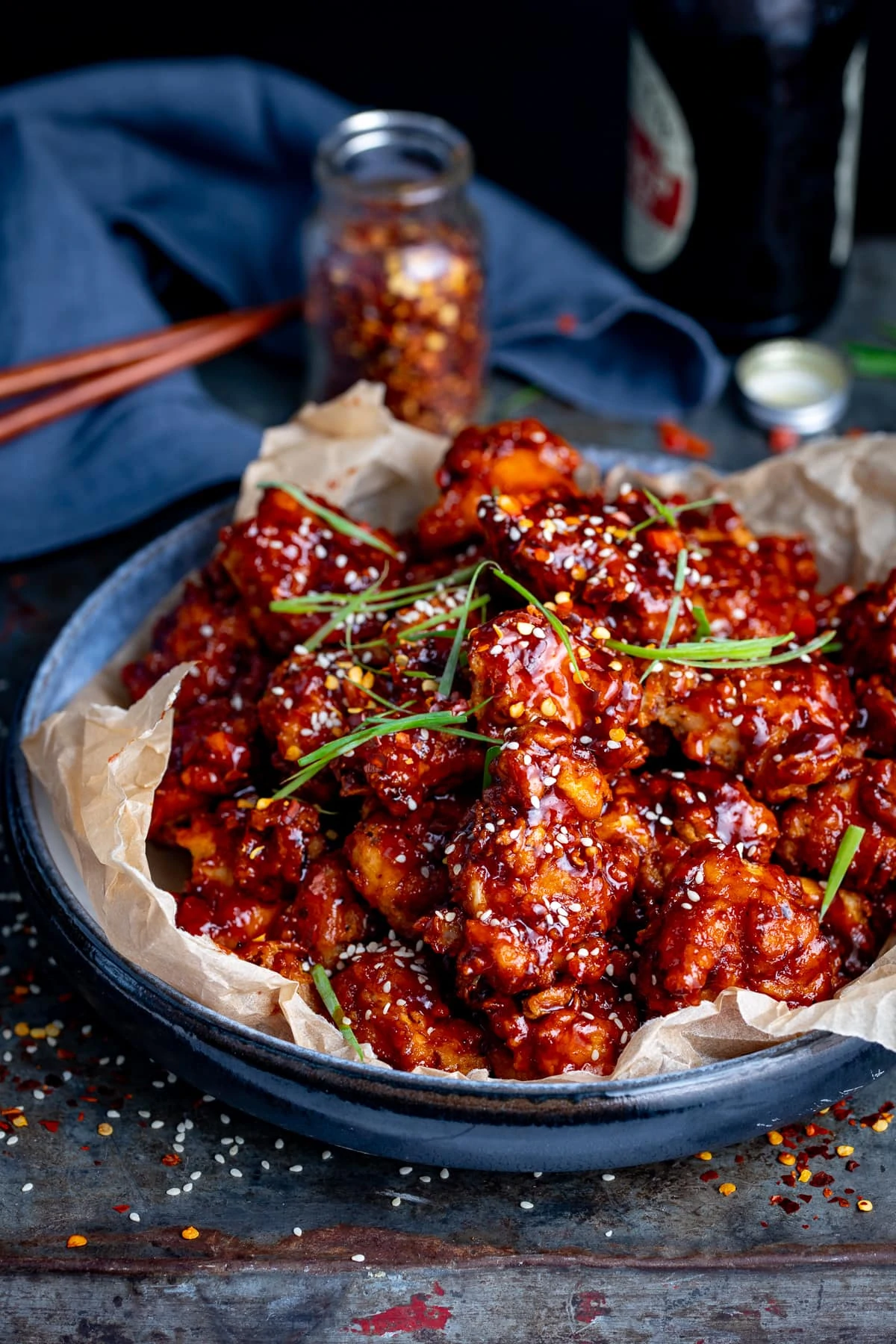 Korean fried chicken in a blue bowl lined with paper. Napkin, chopsticks and jar of chilli flakes in the background.