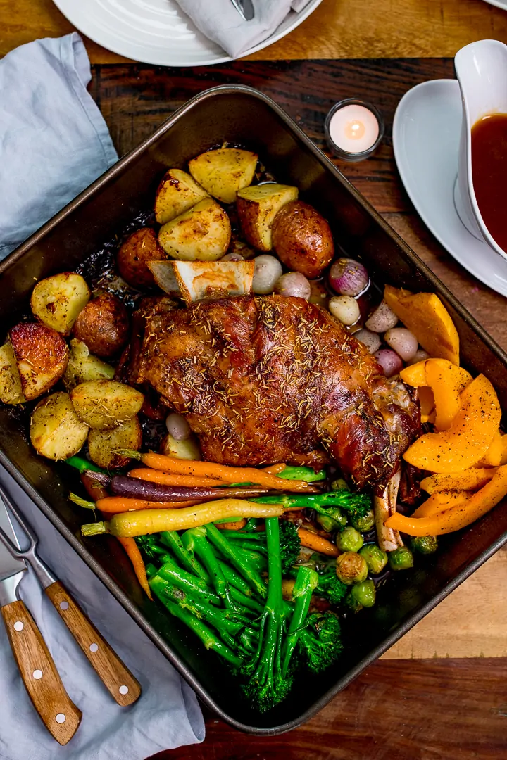 Roast lamb shoulder and vegetables in a roasting tin on a wooden table.