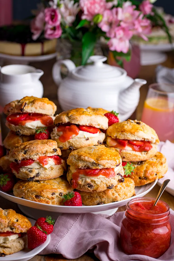 Piled up scones on a white cake stand with jar of rhubarb and strawberry compote. Teapot and milk jug in background.