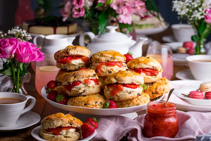 Piled up scones on a white cake stand on a wooden table. Jar of rhubarb and strawberry compote.