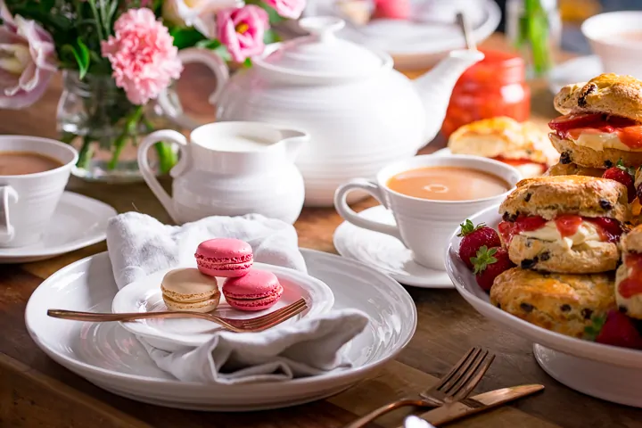 Image of white plates, teapot and milk jug on a table for afternoon tea. Macarons and scones on plates.