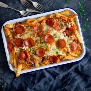 Square image of pizza fries on a white baking tray on a dark blue background with a blue napkin and forks around the tray.