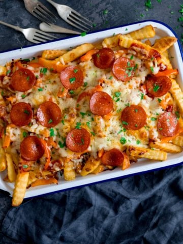 Square image of pizza fries on a white baking tray on a dark blue background with a blue napkin and forks around the tray.