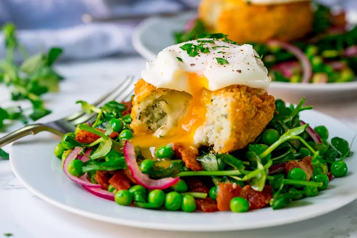 Wide image of pea and bacon salad topped with a fish cake and poached egg - which have been cut into