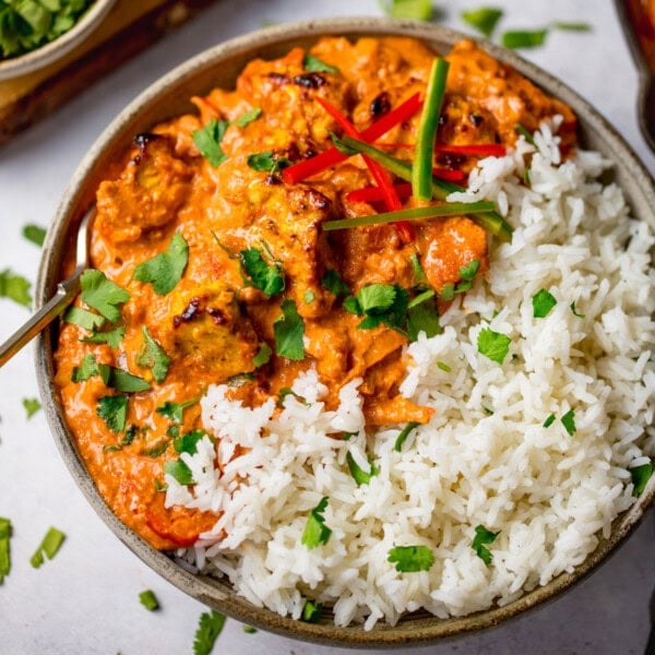 A bowl of chicken tikka masala with white rice, garnished with chopped coriander and sliced chilli peppers. The bowl is on a white surface.