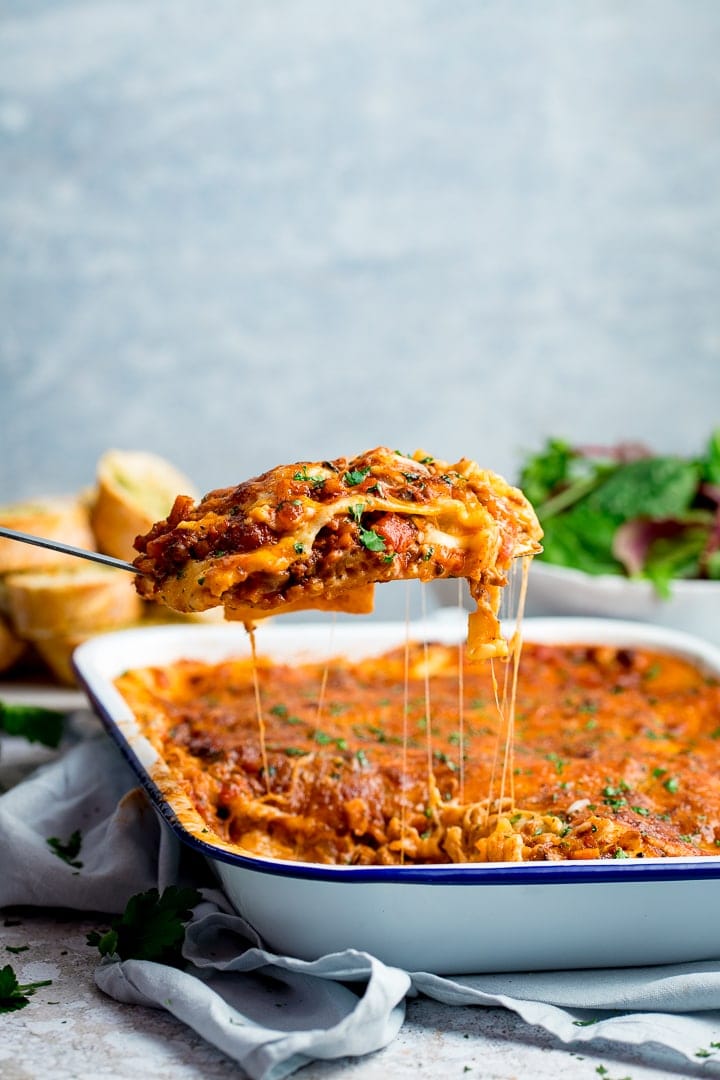 Portion of lasagne being lifted from a dish of lasagne against a light blue background