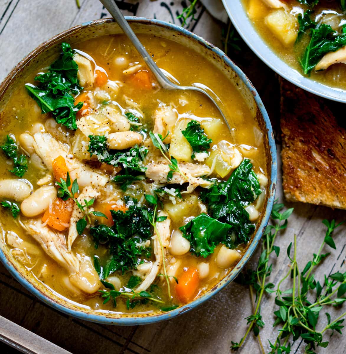 A bowl of Tuscan Chicken Soup with beans, carrots, kale, and herbs, served with a spoon and a slice of toasted bread on the side. The bowl is in a wooden tray and there is a further bowl just in shot.