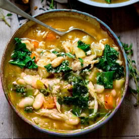 A bowl of Tuscan soup with kale, cannellini beans, carrots, and chicken in a savoury broth, topped with fresh herbs and a spoon inside.