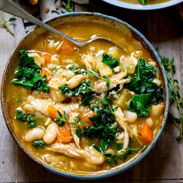 A bowl of Tuscan soup with kale, cannellini beans, carrots, and chicken in a savoury broth, topped with fresh herbs and a spoon inside.