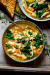 A bowl of hearty Tuscan Chicken Soup with shredded chicken, white beans, carrots, kale, and fresh herbs, served with toasted bread on the side. The bowl is in a wooden tray and there is a further bowl just in shot.
