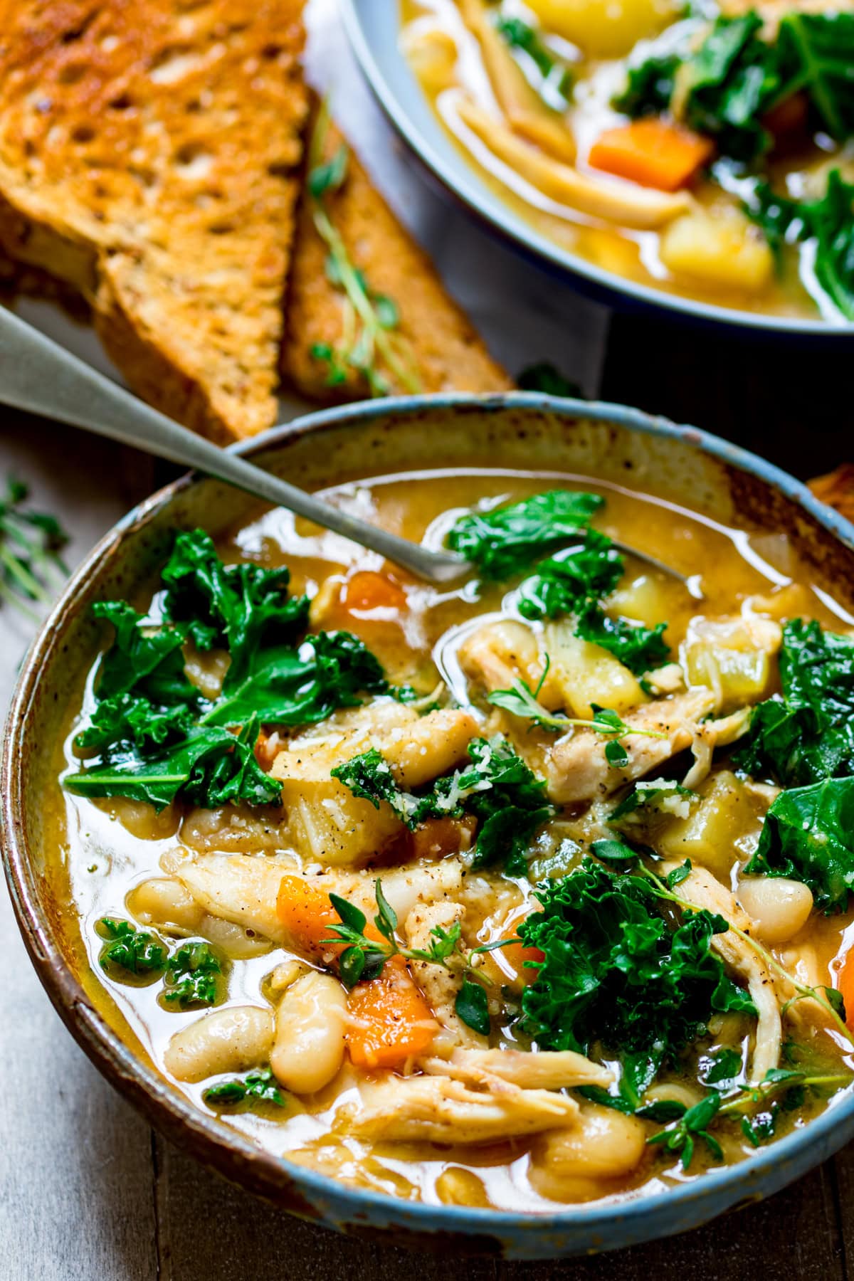 A bowl of hearty Tuscan Chicken Soup with shredded chicken, white beans, kale, carrots, and potatoes, served with toasted bread on the side. The bowl is in a wooden tray and there is a further bowl just in shot.