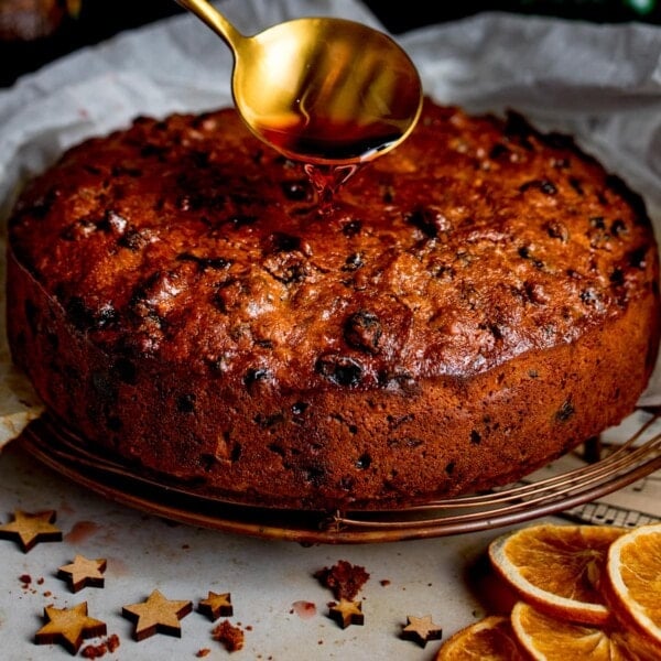 Cherry brandy being spooned onto a christmas cake with orange slices and wooden stars in the foreground.