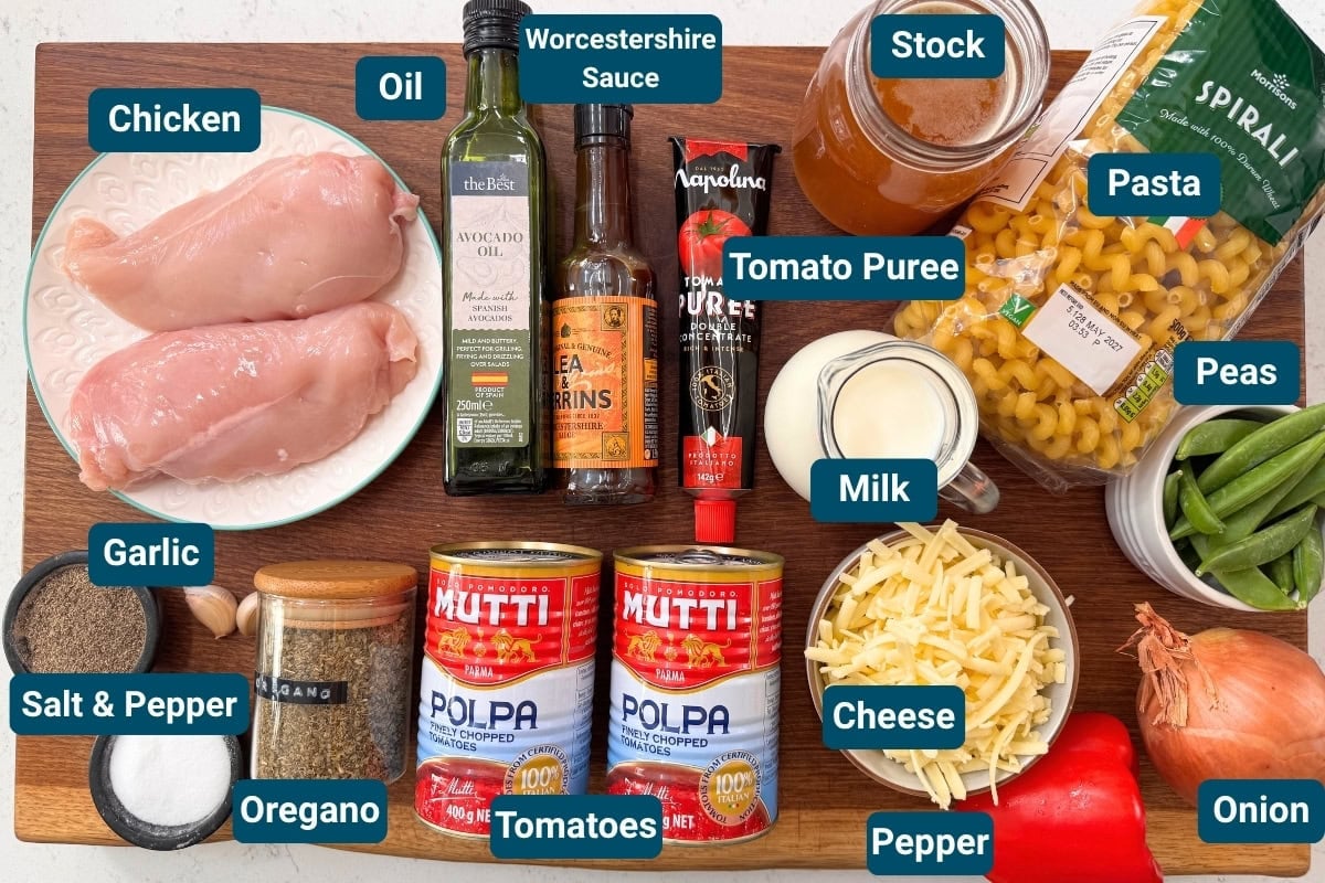 A wooden board with raw chicken fillets, tinned tomatoes, cheese, pasta, milk, tomato purée, oil, peas, stock, garlic, onion, spices and sauces for a one pot chicken pasta.