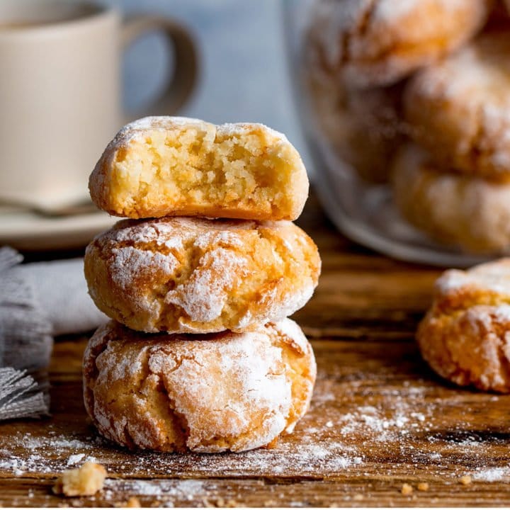 Italian Amaretti cookies in a pile on a wooden board with a cup in the background.