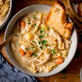 A bowl of homemade chicken noodle soup with pasta, carrots, and potatoes, garnished with parsley and served with toasted buttered bread. The soup bowl in on a wooden table.
