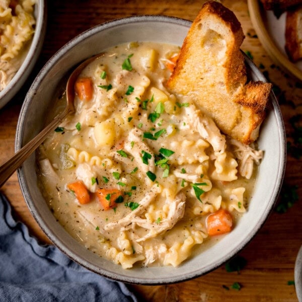 A bowl of homemade chicken noodle soup with pasta, carrots, and potatoes, garnished with parsley and served with toasted buttered bread. The soup bowl in on a wooden table.