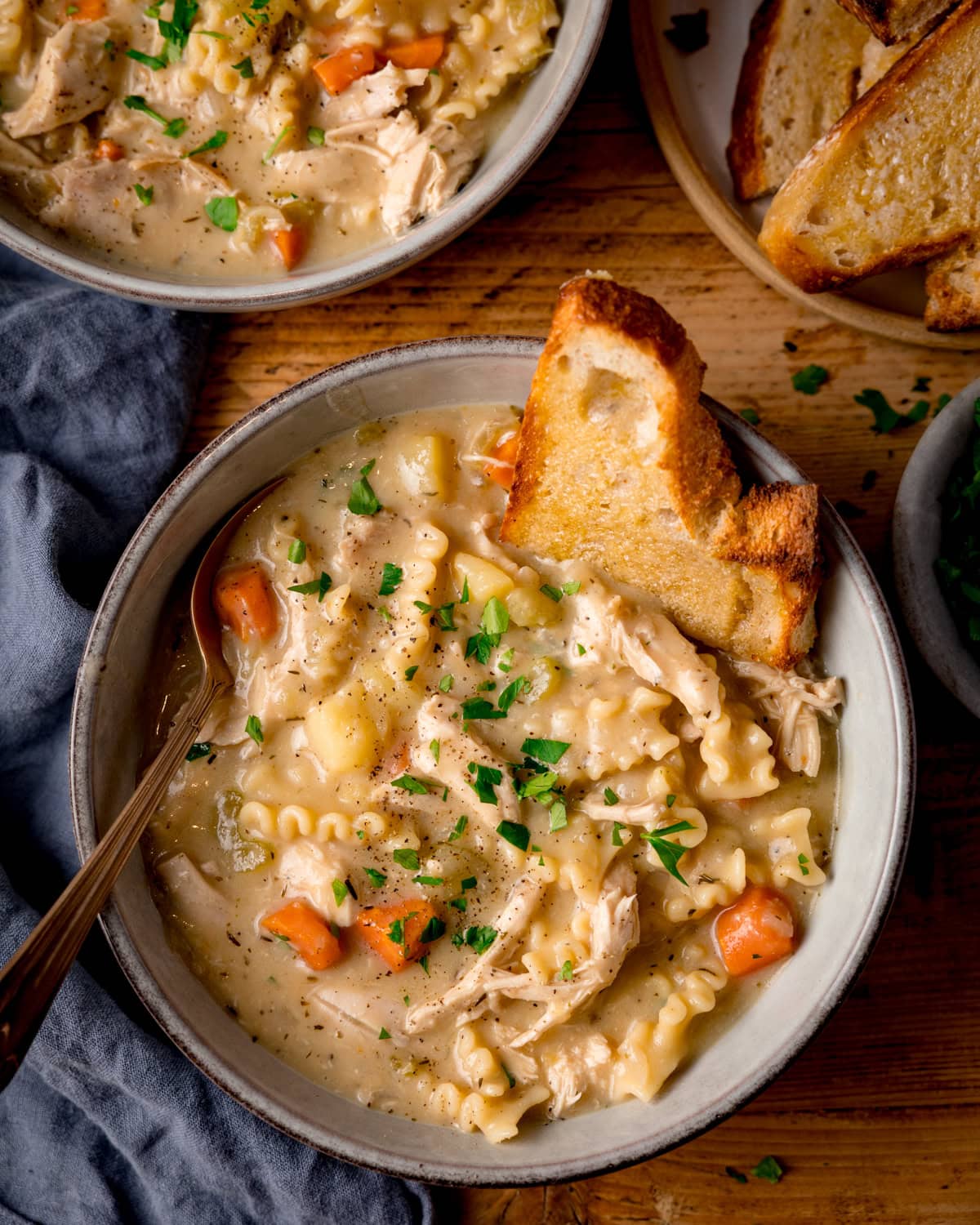 A bowl of homemade chicken noodle soup with pasta, carrots, and potatoes, garnished with parsley and served with toasted buttered bread. The soup bowl in on a wooden table with a further bowl just in shot at the top of the frame.