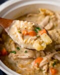 A close-up of a wooden spoon holding creamy Chicken Noodle Soup with carrots, potatoes, noodles, and herbs over a white soup pot.