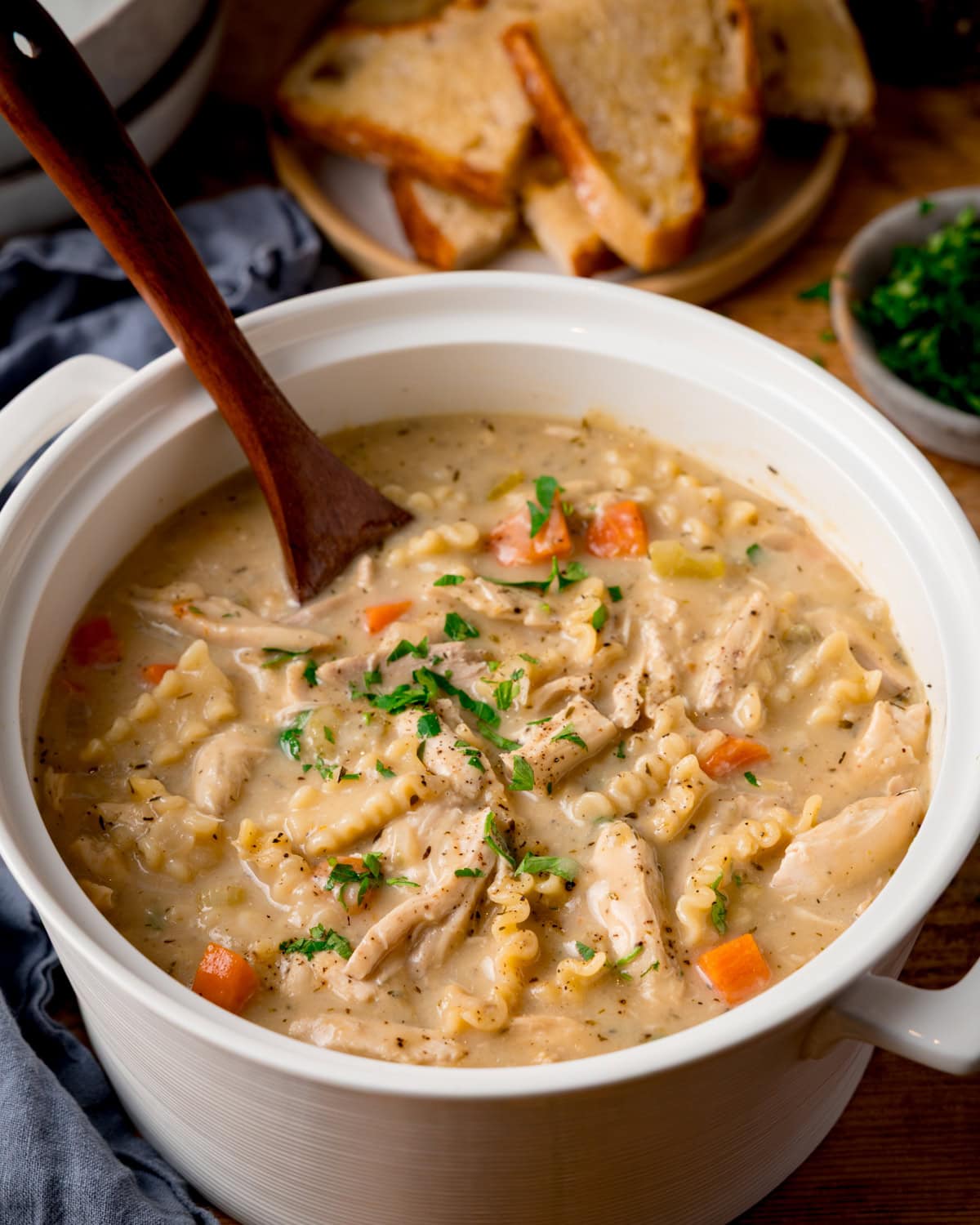 A pot of creamy chicken noodle soup with carrots and herbs. There is a wooden spoon sticking out. A place of buttered toast pieces is in the background alongside a small bowl of chopped parsley and a blue napkin.