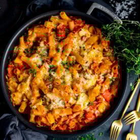 Baked rigatoni pasta with melted cheese, tomato sauce, turkey and ham in a black dish, topped with parsley. Gold forks and fresh thyme nearby.