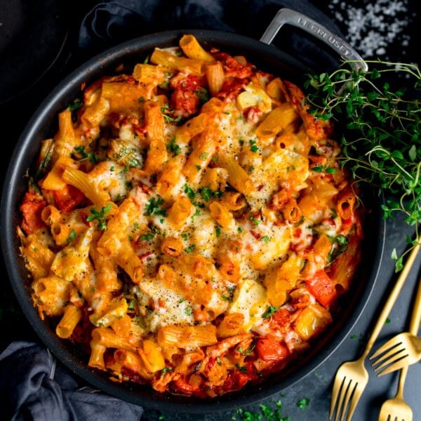Baked rigatoni pasta with melted cheese, tomato sauce, turkey and ham in a black dish, topped with parsley. Gold forks and fresh thyme nearby.