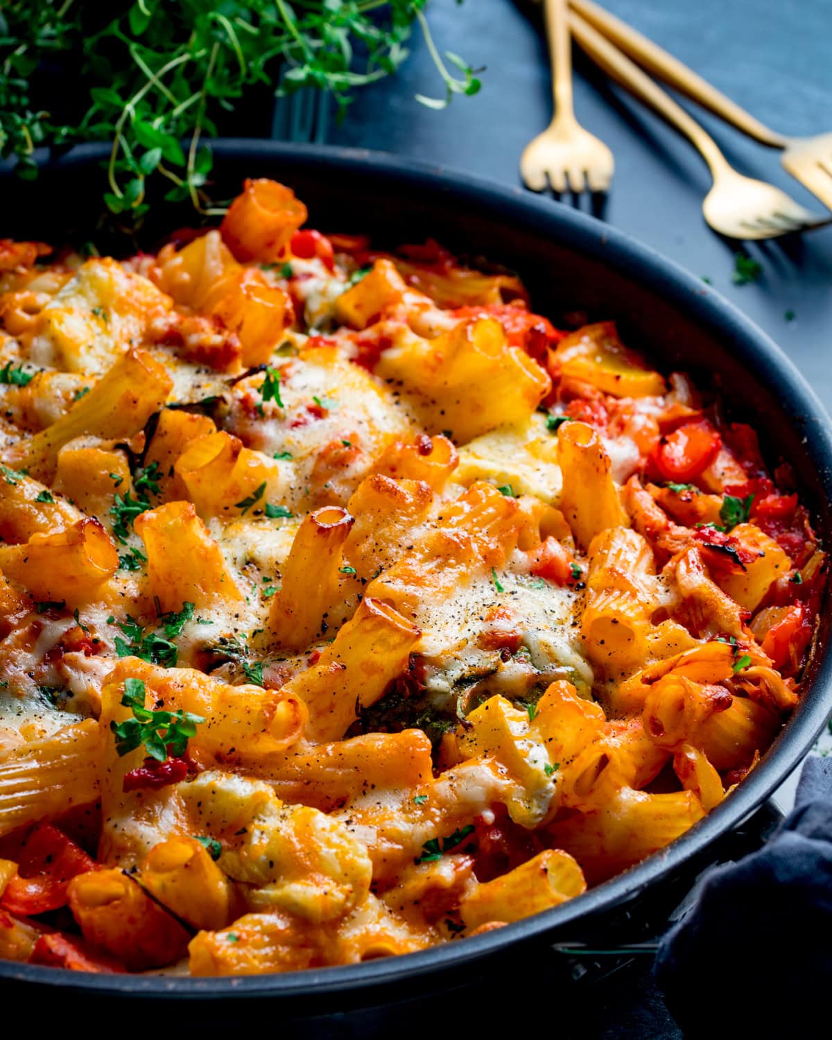 Baked rigatoni pasta with melted cheese, tomato sauce, turkey and ham in a black pan. Served with gold forks and fresh parsley in the background.