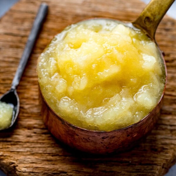 A metal dish filled with chunky apple sauce for pork sits on a wooden surface, with a spoon containing a small amount of apple sauce beside it.