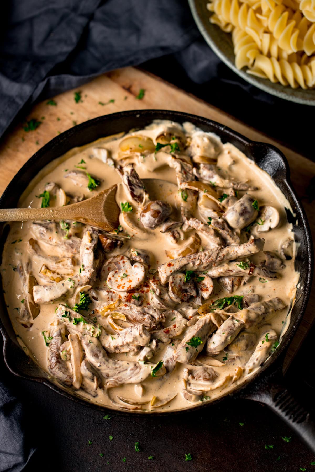A cast iron frying pan filled with creamy mushroom and beef stroganoff, garnished with parsley and paprika. A wooden spoon rests in the pan. There is a bowl of pasta at the top of the frame.