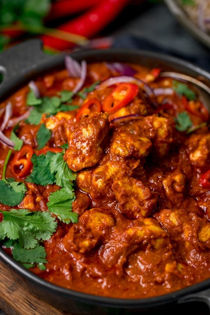 Close up of chicken madras in a dark bowl. The madras is topped with sliced coriander, red onion and red chillies.